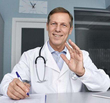 Médico sorrindo em consultório enquanto escreve e cumprimenta paciente.