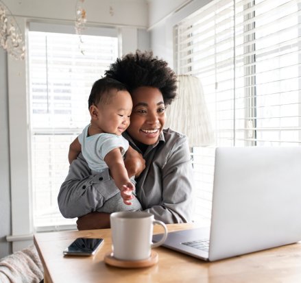Mãe com bebê no colo usando notebook em casa para atendimento online.