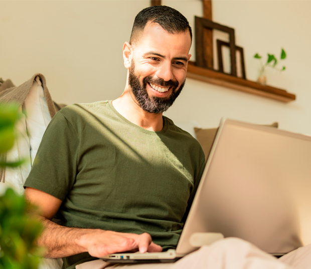 Homem sorrindo usando notebook em casa, sentado confortavelmente em ambiente bem iluminado.